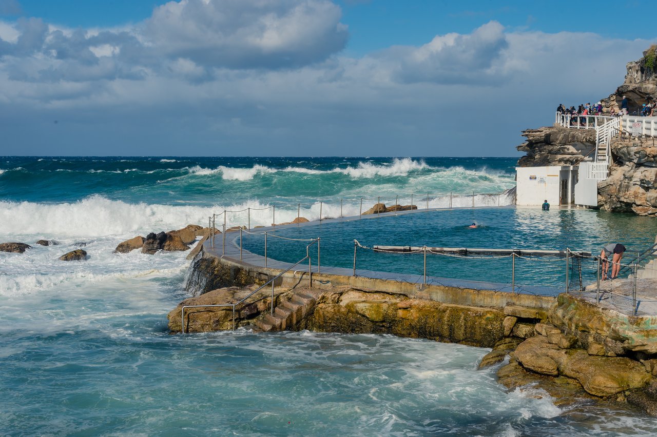 An ocean pool with swimmers, waves crashing nearby, and people watching from a rocky cliffside walkway.