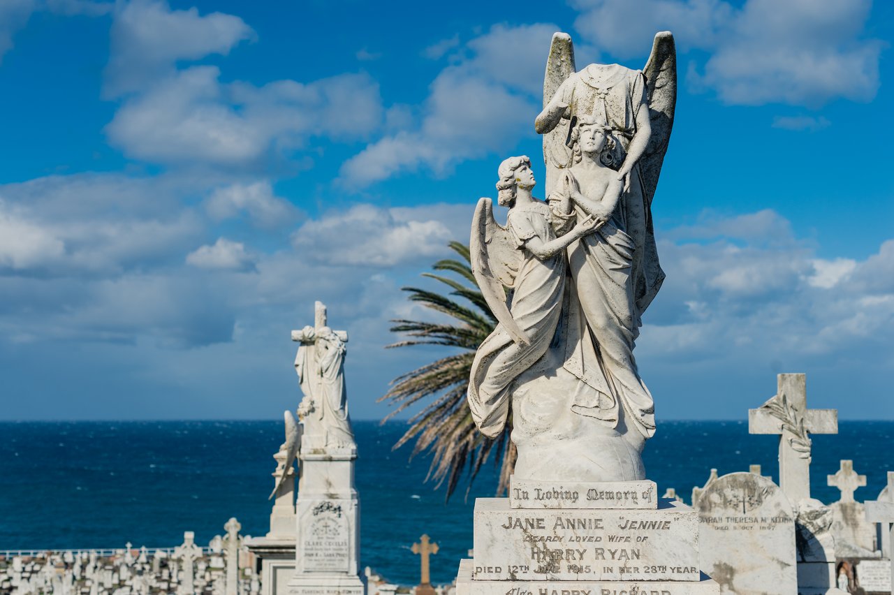 A weathered cemetery statue of angels and a headless figure stands atop a gravestone under a blue sky.