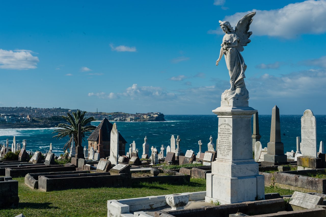 A cemetery overlooking the ocean with a large angel statue on a tombstone under a clear blue sky.