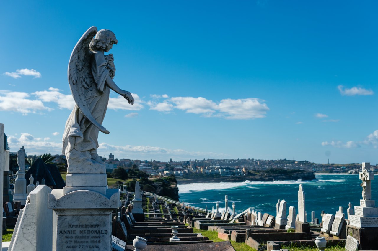 A stone angel statue stands on a grave in a cemetery overlooking the ocean.