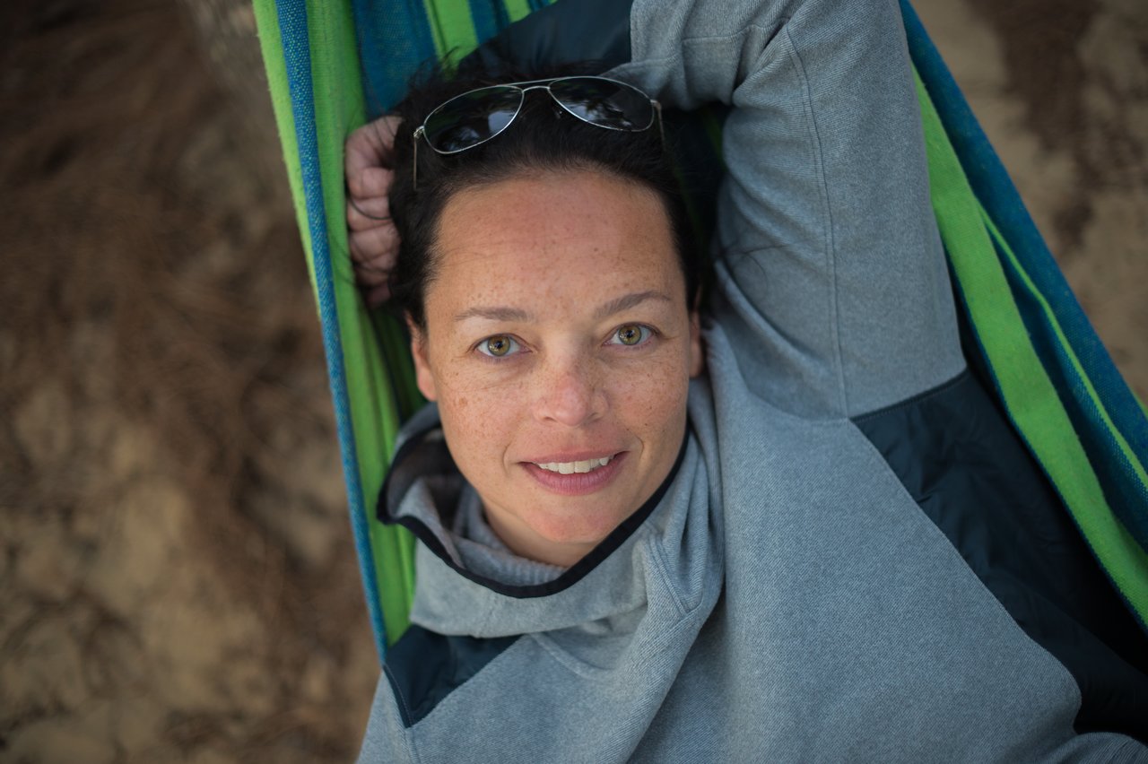 A woman in a gray hoodie lies in a hammock, smiling and looking at the camera.