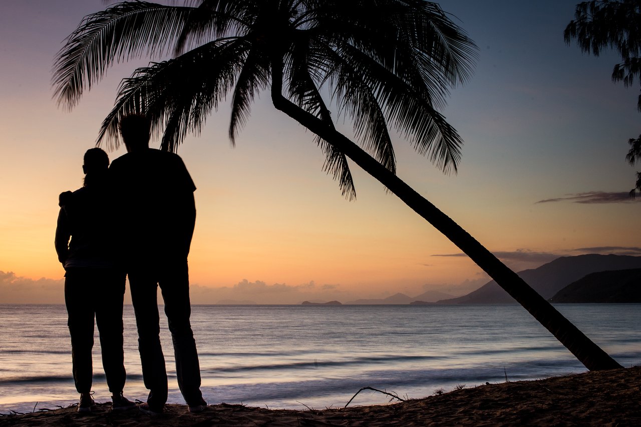 Two people stand close together on a beach, looking at the ocean during sunrise.