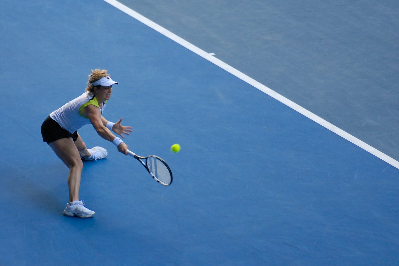 A tennis player stretches to return a shot during a match on a blue hard court.
