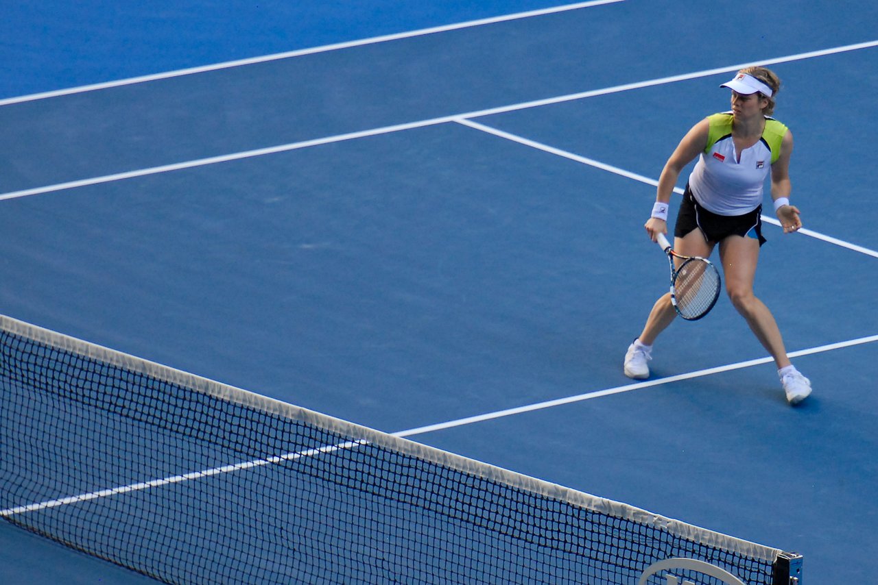A tennis player prepares to return a shot during a match on a blue hard court.