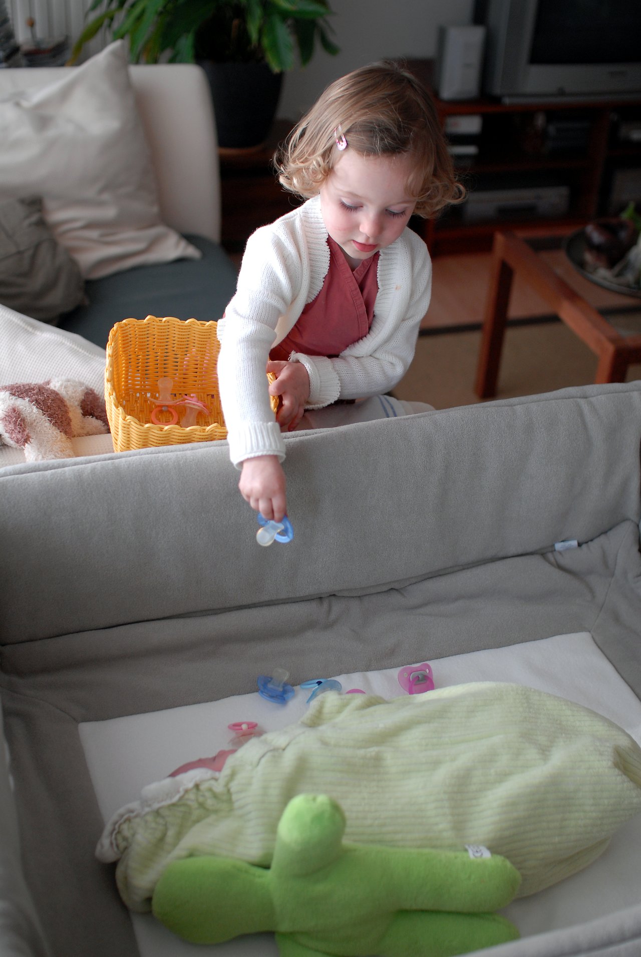 A young child places pacifiers into a crib where a baby is sleeping, holding a basket with more pacifiers.