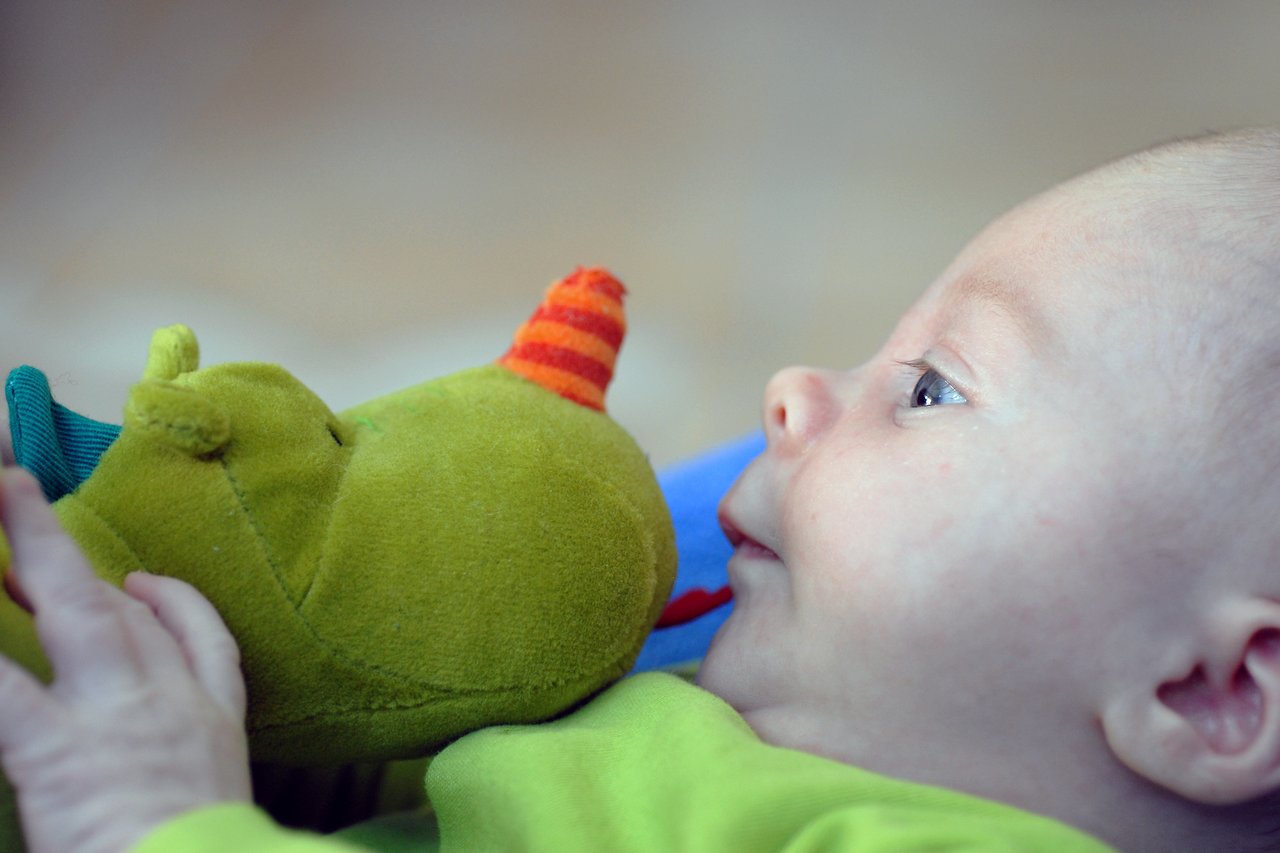 A baby in a green outfit holds a plush dragon toy and playfully puts its nose in their mouth.