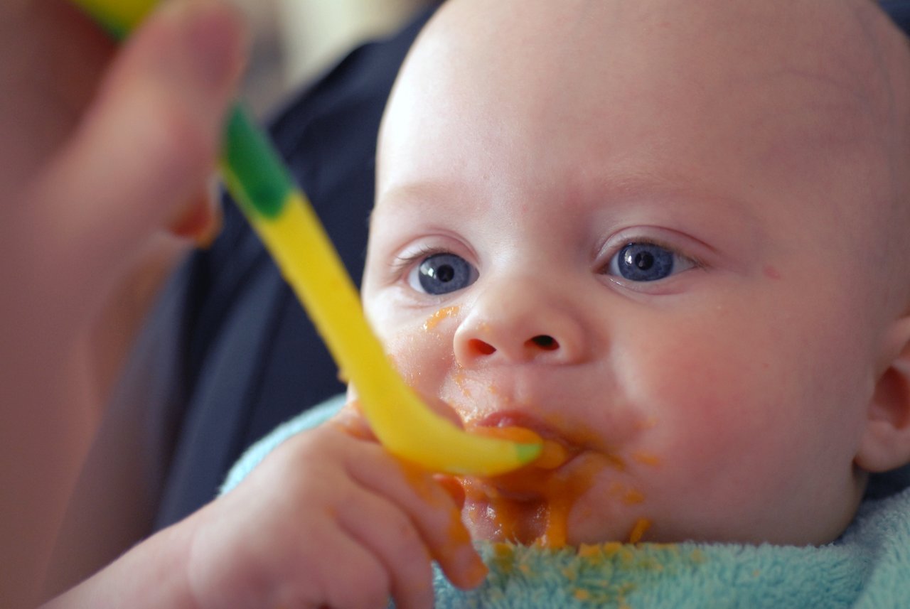 A baby with a blue bib eats mashed vegetables from a spoon, with some food smeared on their face.