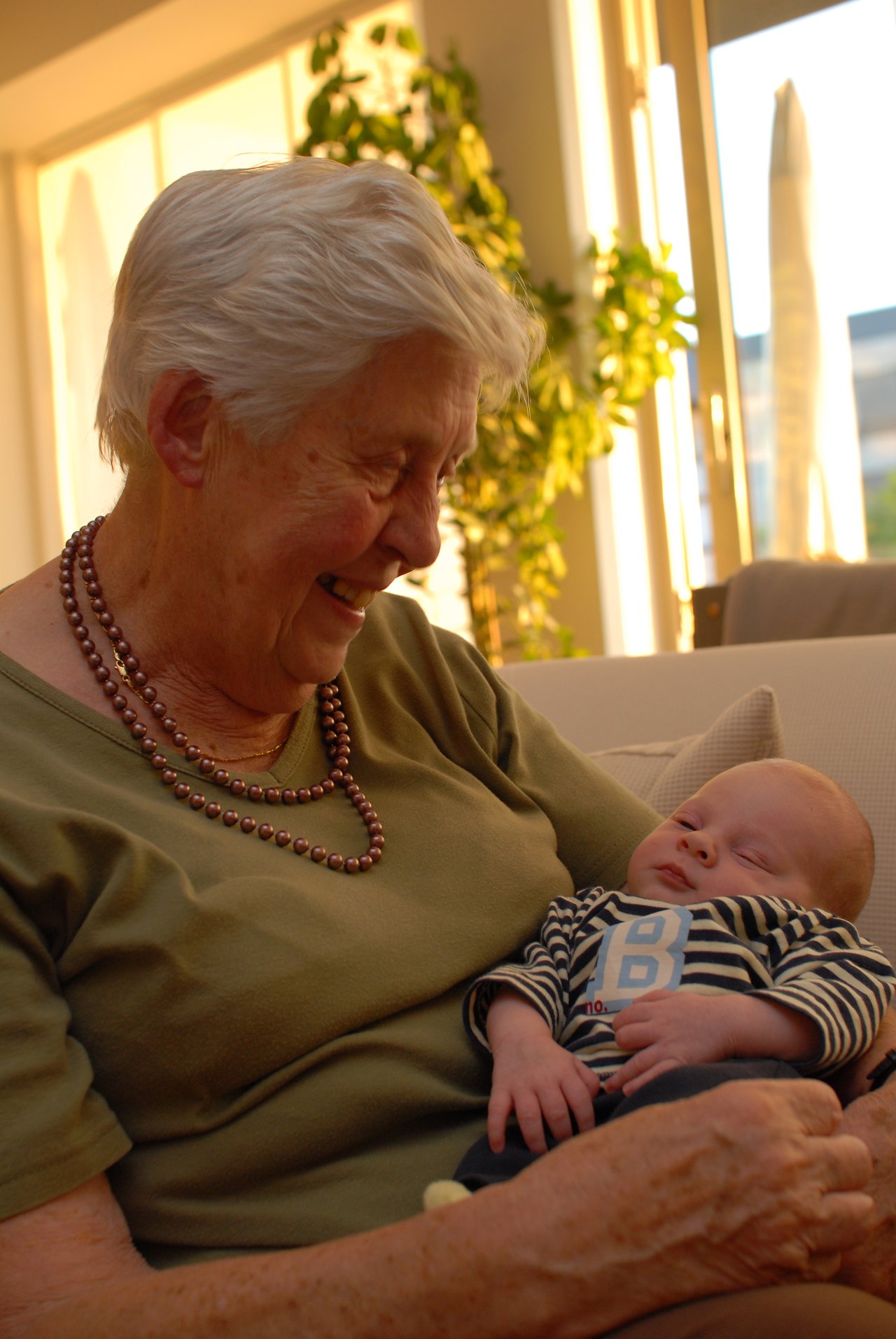 An elderly woman smiles while holding a sleeping baby in her arms.