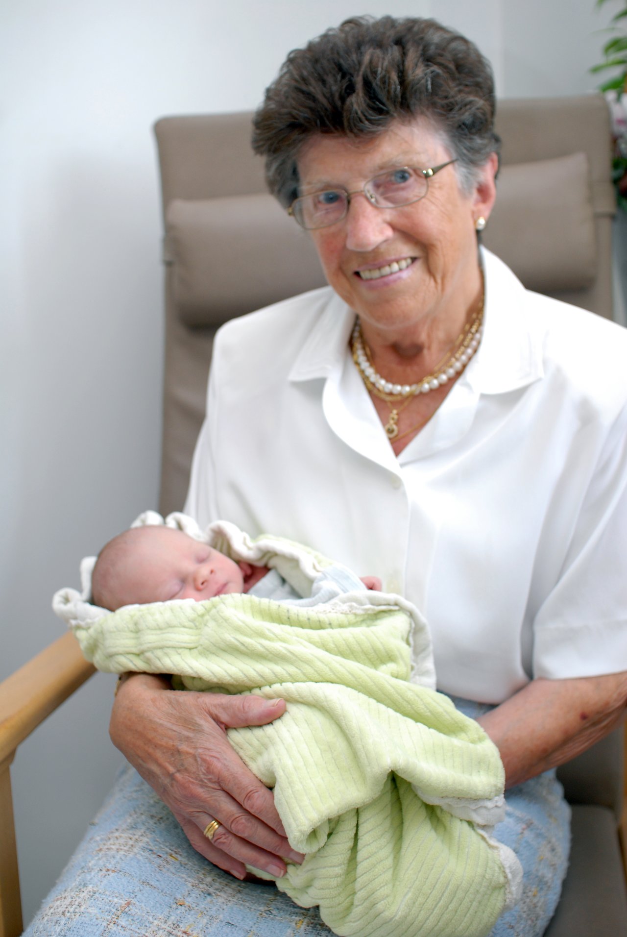 An elderly woman wearing glasses and a white blouse holds a sleeping newborn wrapped in a soft green blanket.