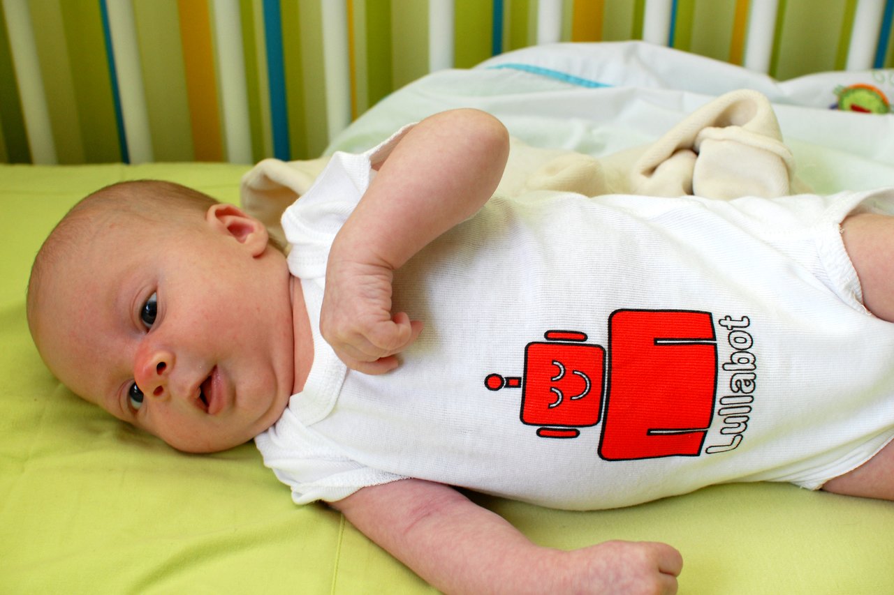 A baby lies on a green surface, wearing a white onesie with a red robot design and the word "Lullabot.