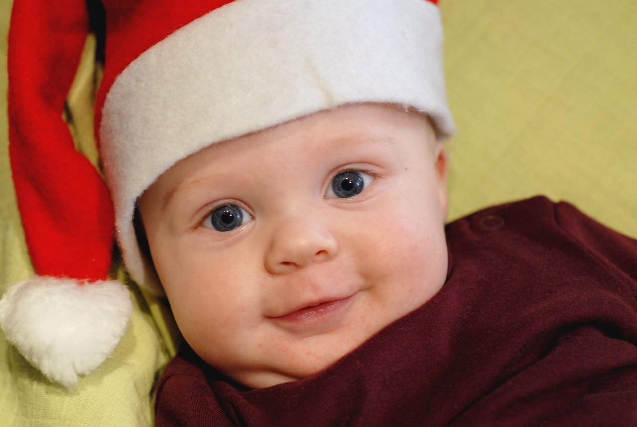 A baby wearing a Santa hat smiles while lying down on a soft surface.