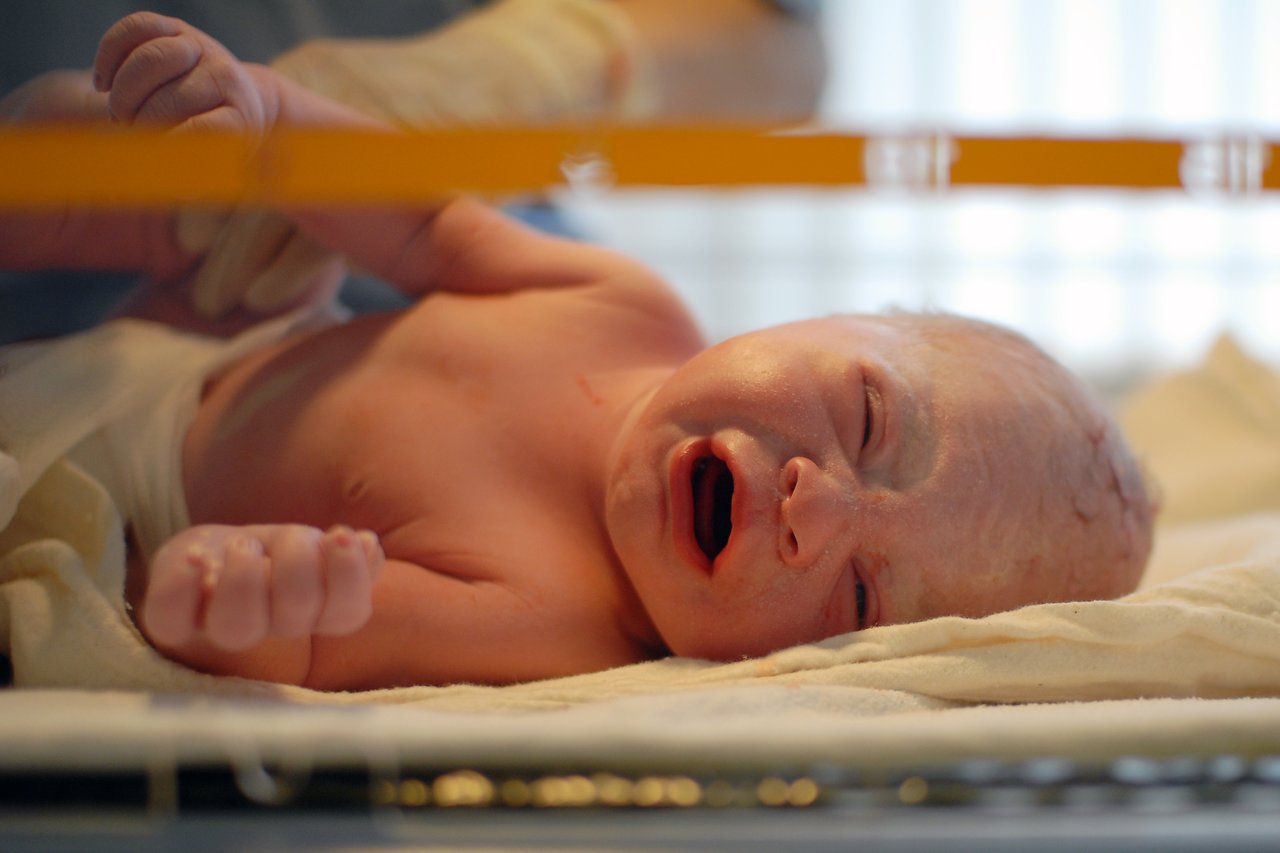 A newborn baby lies on a hospital bed, crying, while a gloved hand supports its arm.