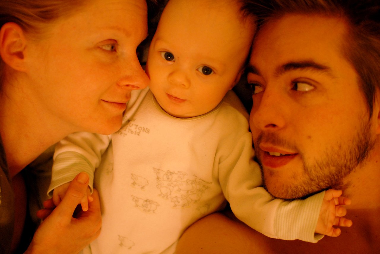 A baby lies between two parents on a bed, holding their hands and looking at the camera.