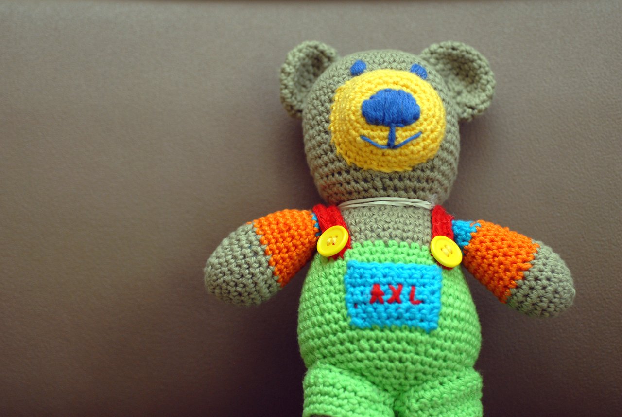 A handmade crocheted teddy bear with colorful clothing sits against a brown background.