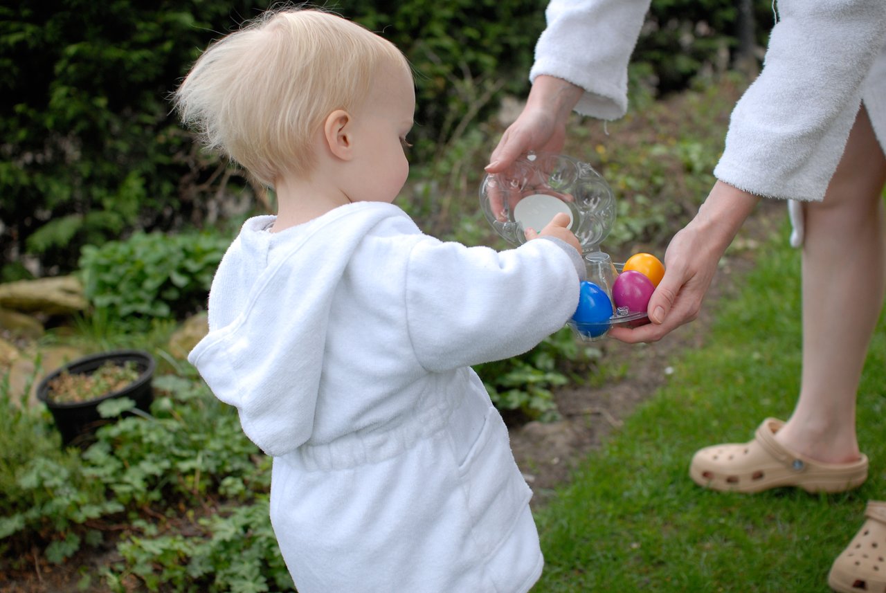 A child in a white hoodie reaches for colorful Easter eggs held by an adult in a garden.