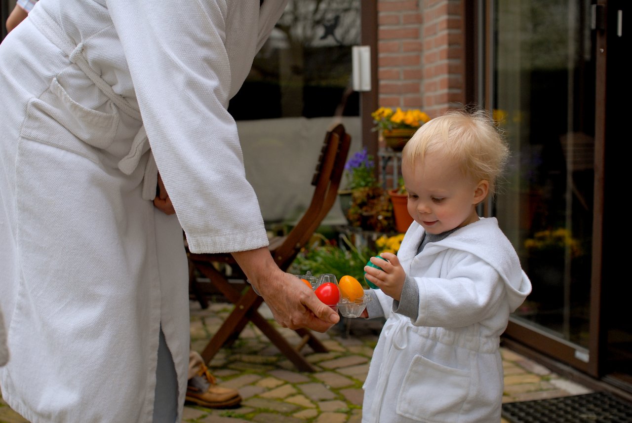 A child in a white robe smiles while receiving colorful Easter eggs from an adult in a matching robe.