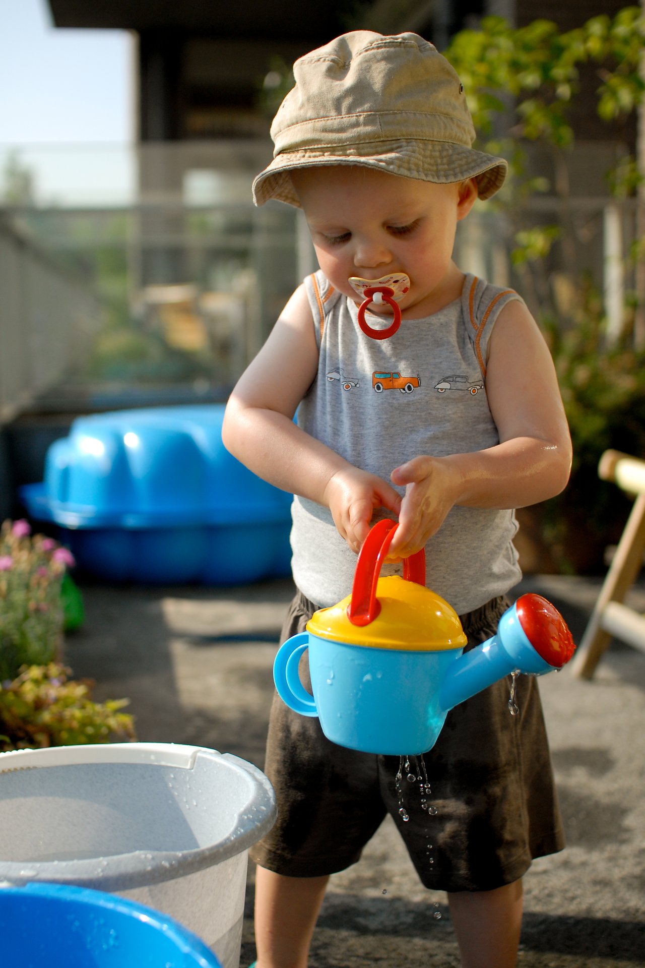 A young child wearing a hat and pacifier pours water from a toy watering can on a terrace.