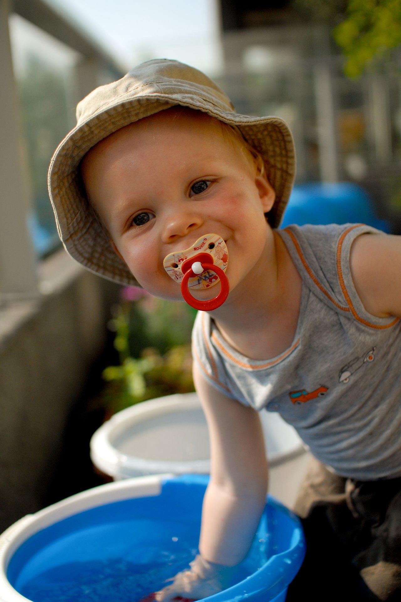 A young child wearing a hat and pacifier plays with water in a blue bucket on a terrace.