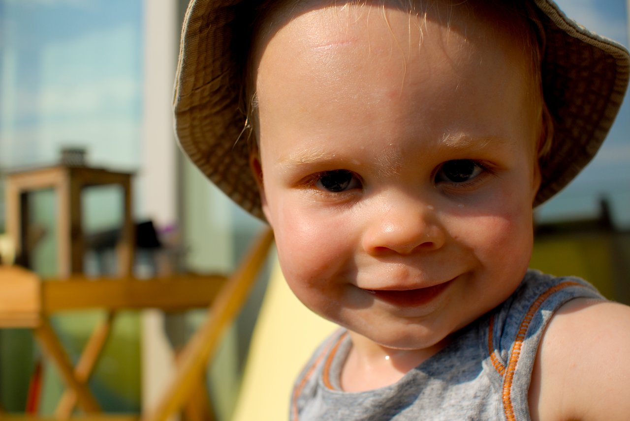 A smiling toddler wearing a sun hat plays on a terrace, looking directly at the camera.