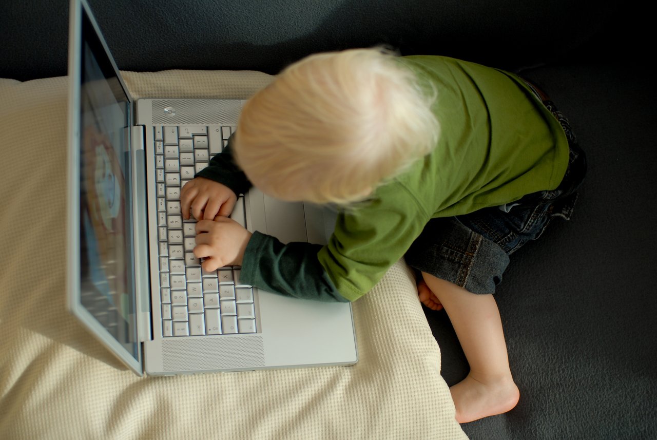 A young child with blonde hair types on a laptop while sitting on a couch, focused on the screen.