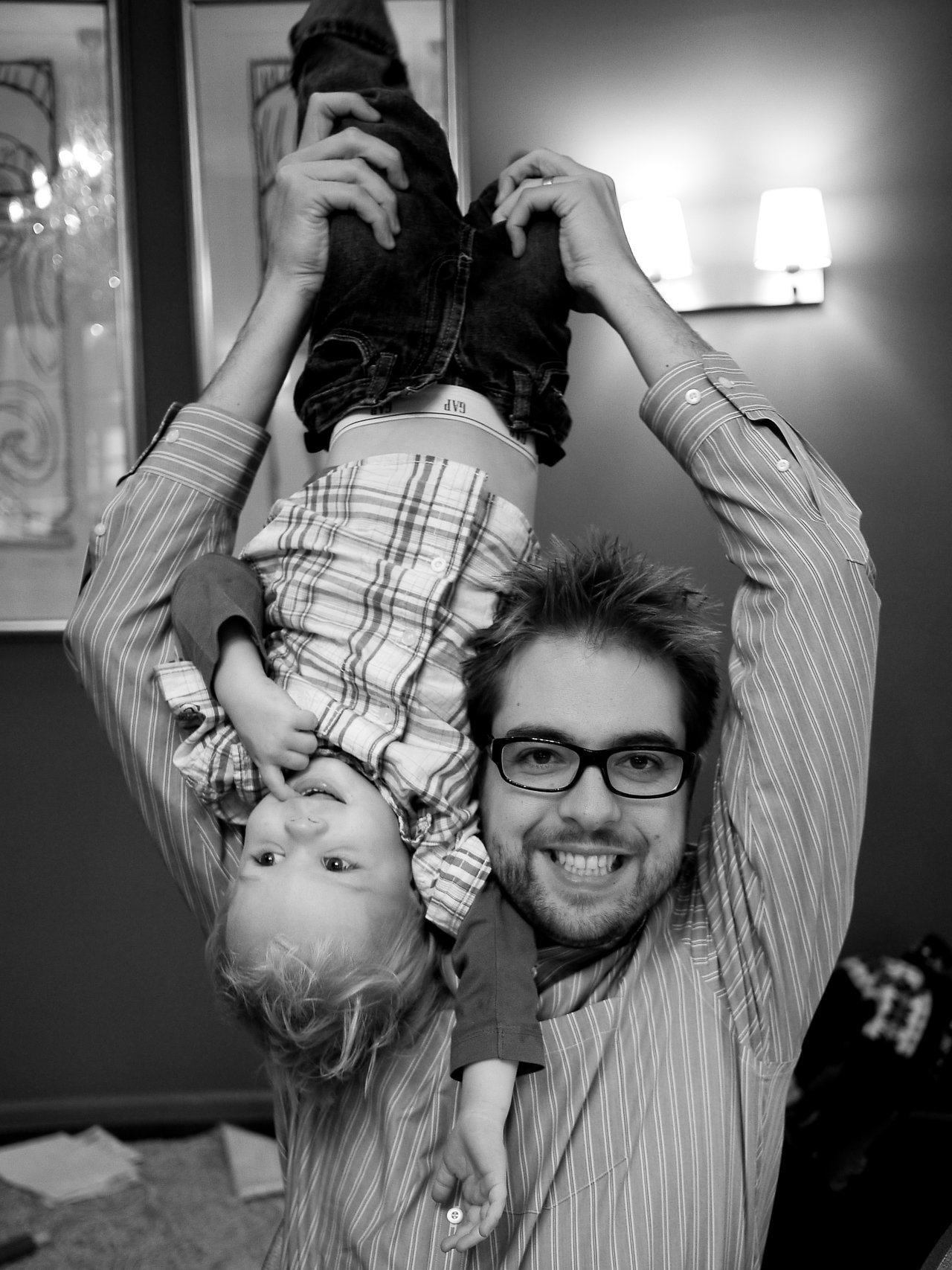 A man smiles while playfully holding a young child upside down by the legs indoors.