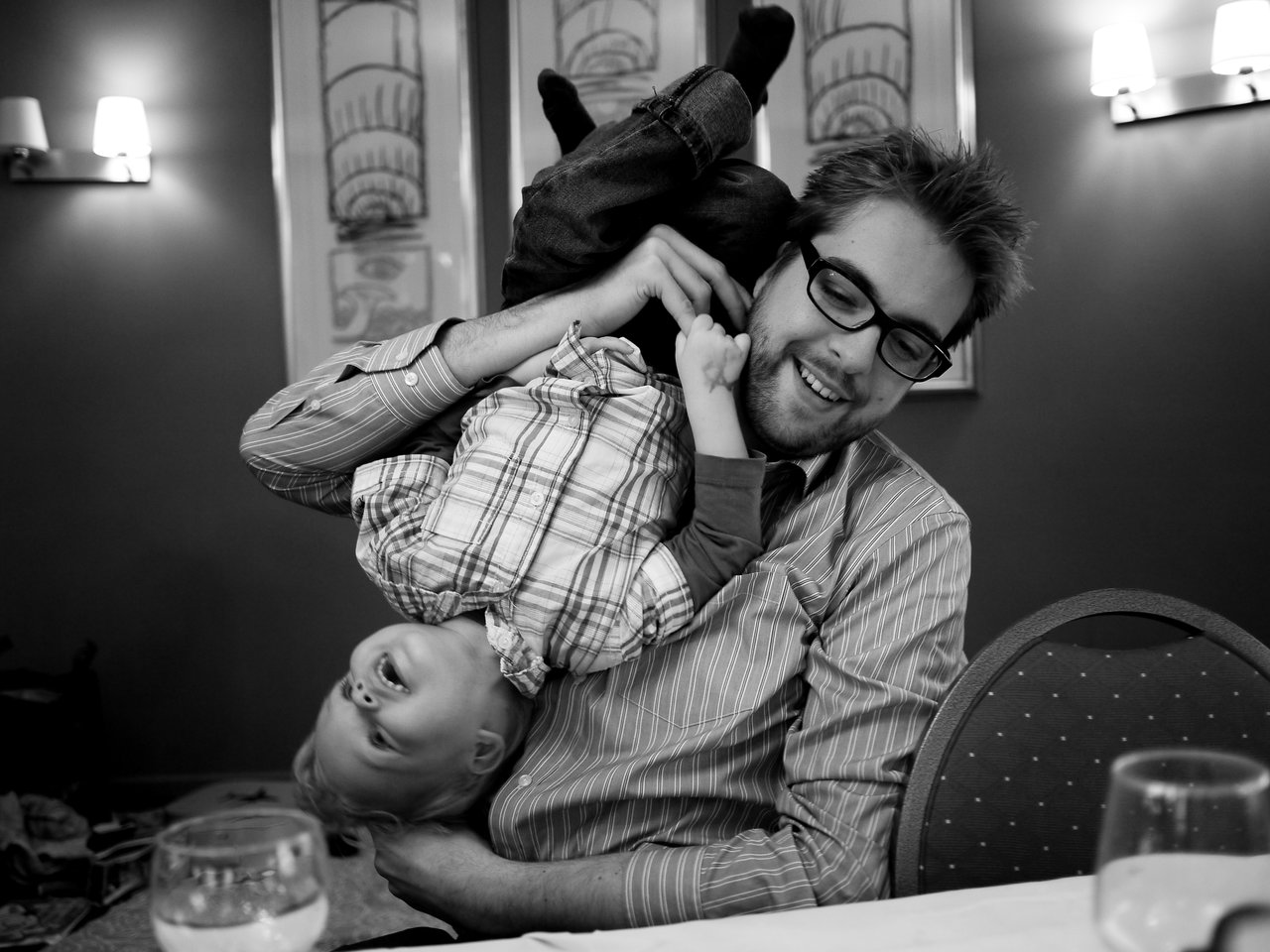 A man wearing glasses smiles while playfully holding a young child upside down over his shoulder.