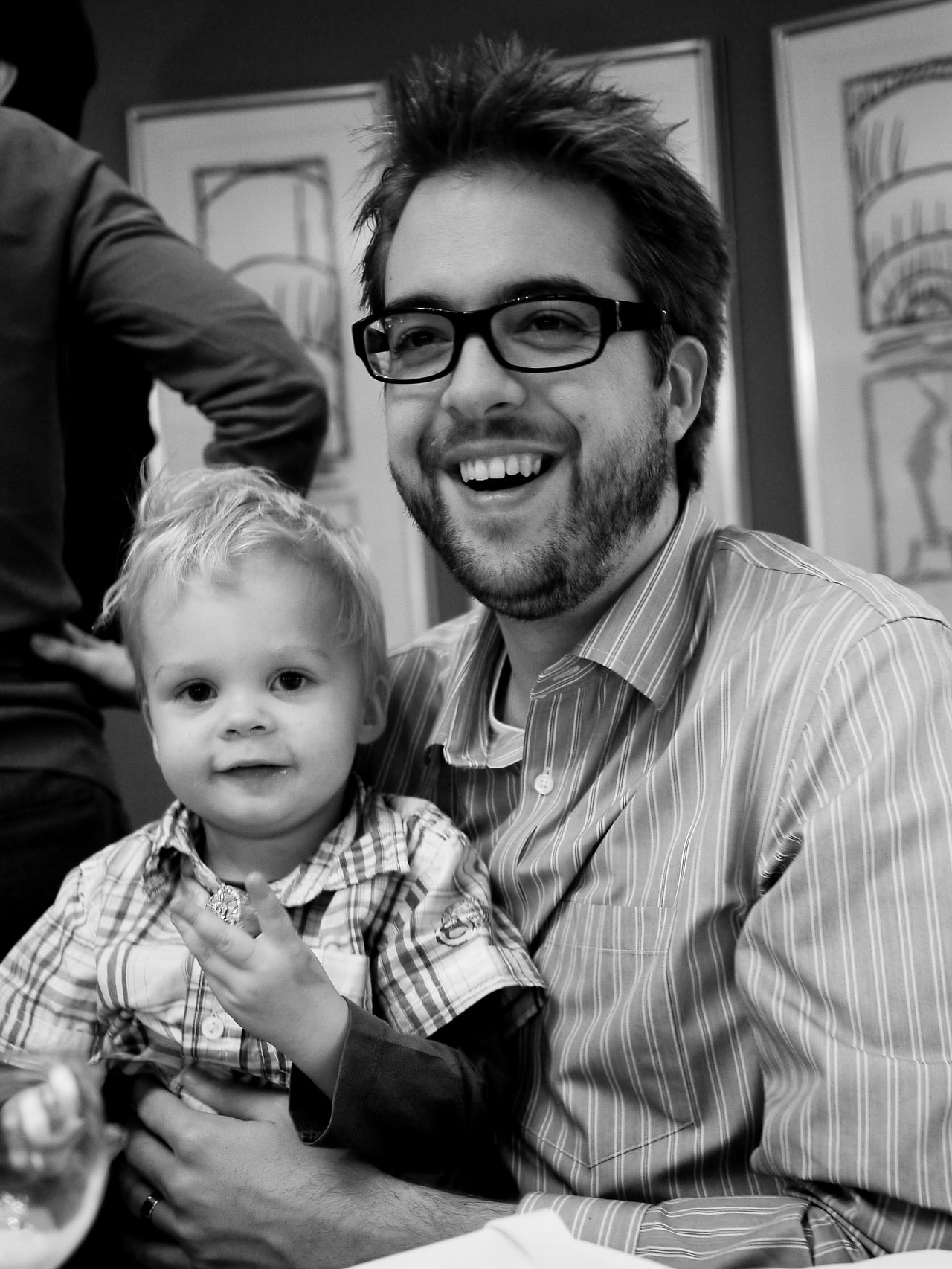A man with glasses and a beard smiles while holding a young child in his arms at a gathering.