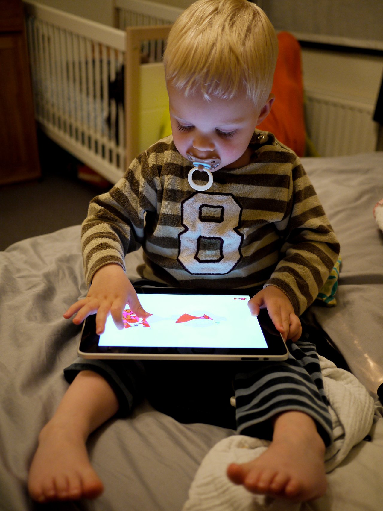 A young child with a pacifier sits on a bed, using a tablet with both hands.
