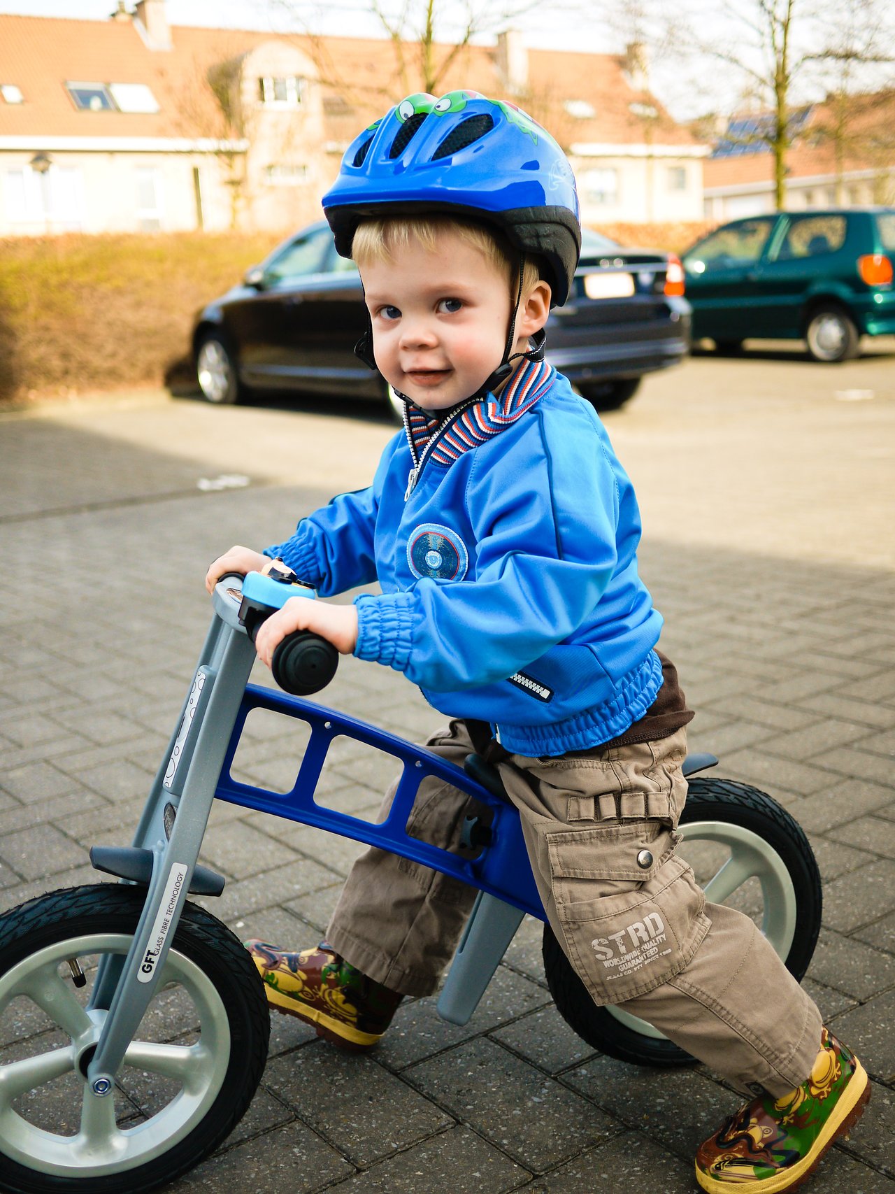 A young child wearing a blue helmet rides a balance bike on a paved surface, looking ahead confidently.