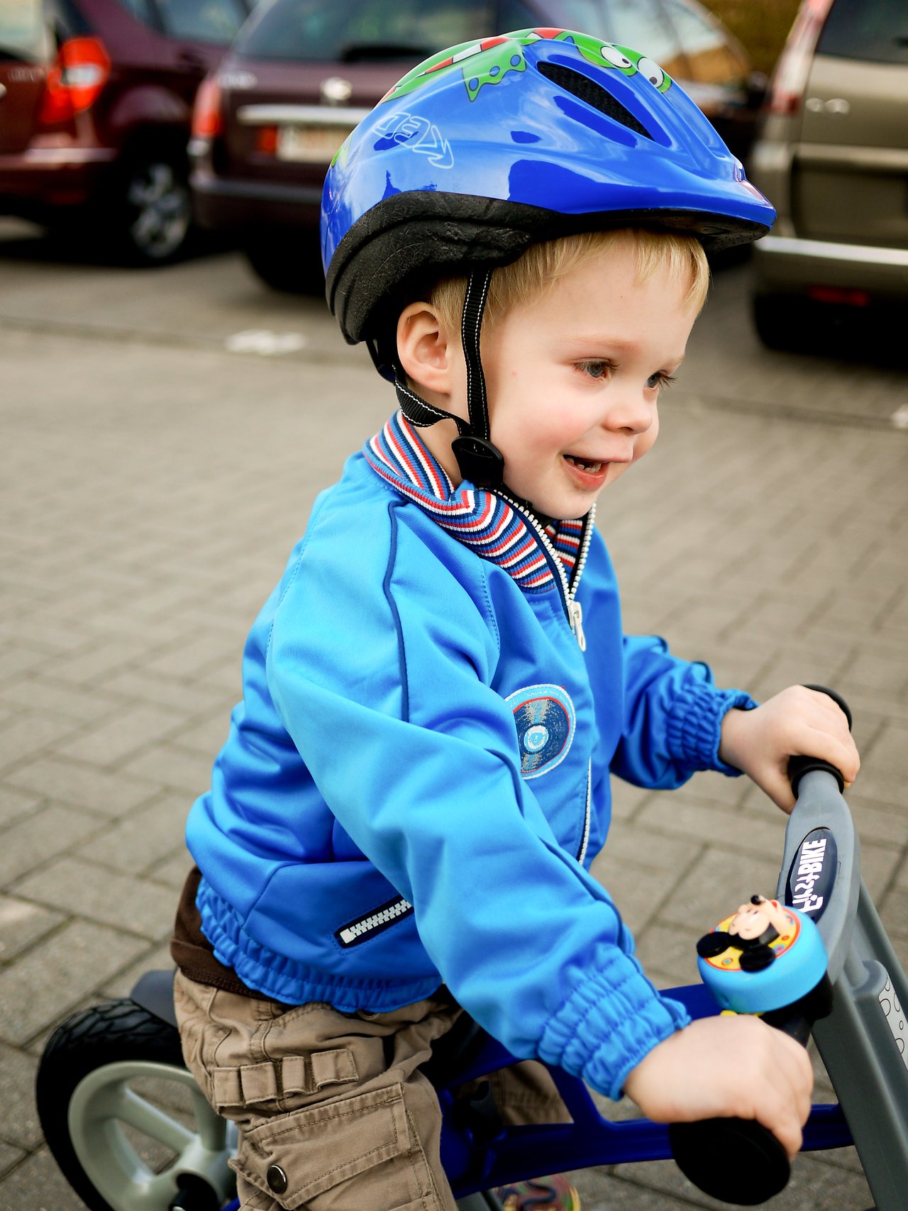 A young child wearing a blue helmet and jacket rides a small bike, gripping the handlebars and smiling.