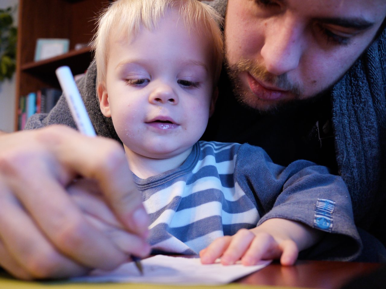 A young child holds a pen while an adult guides their hand, helping them learn to write on paper.