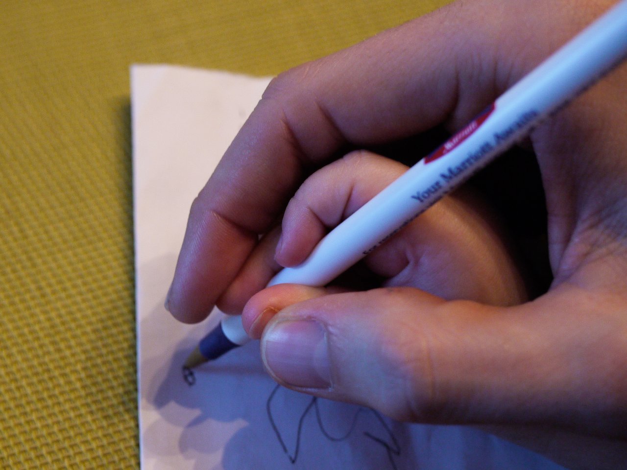 An adult hand guides a child's small hand in holding a pen and writing on a piece of paper.