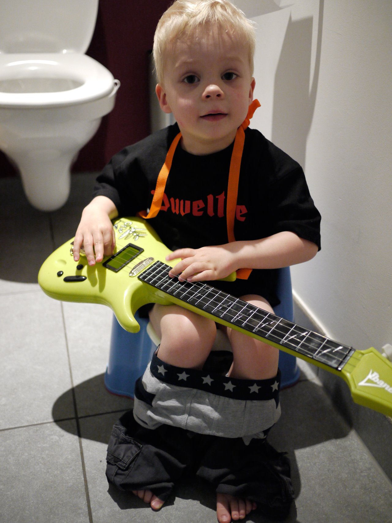 A young child sits on a potty with pants down, holding a green toy guitar and looking at the camera.