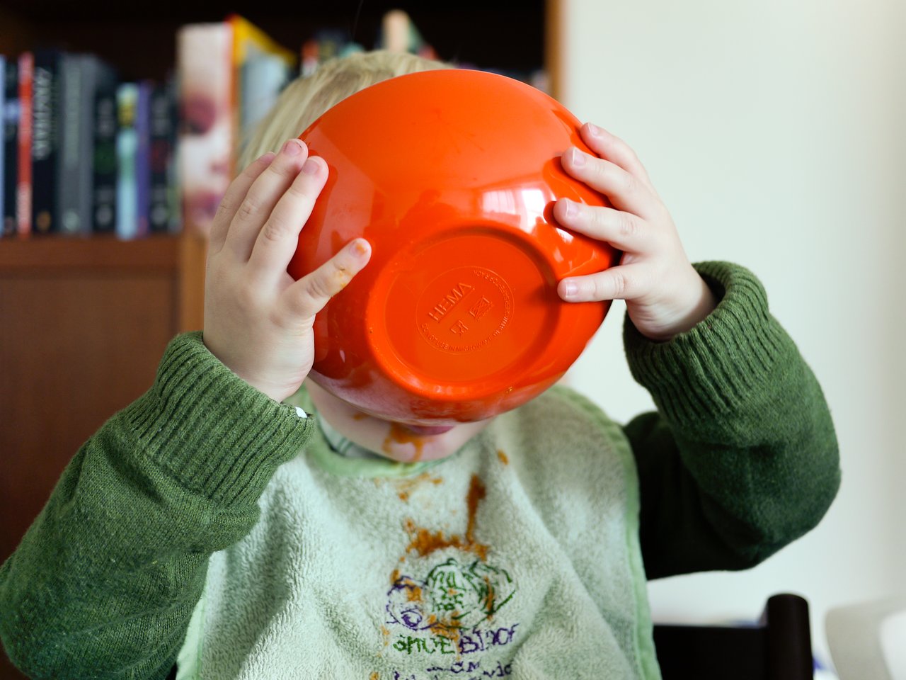 A child in a green sweater drinks soup directly from an orange bowl, with soup stains on their face and bib.