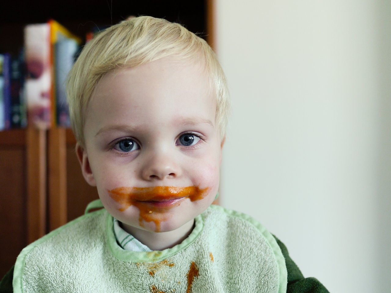 A young child with a messy face and bib has soup smeared around their mouth, forming a "mustache.