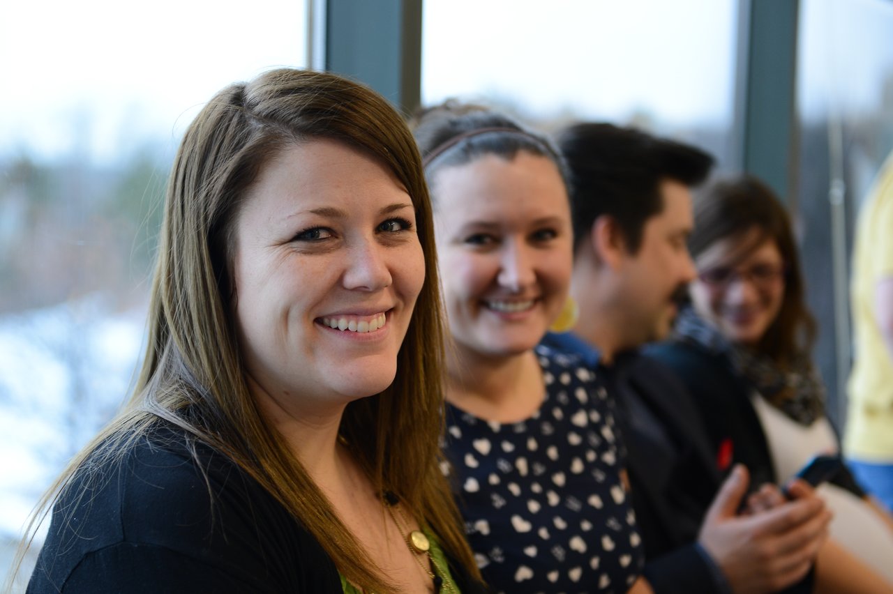 Two smiling women sit together at a baby shower, with other guests chatting in the background.