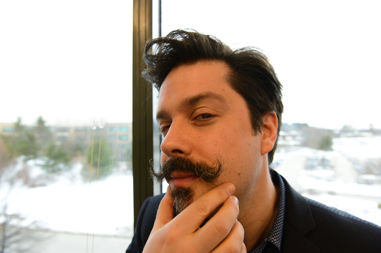 A man with a mustache poses playfully, touching his chin and looking at the camera.