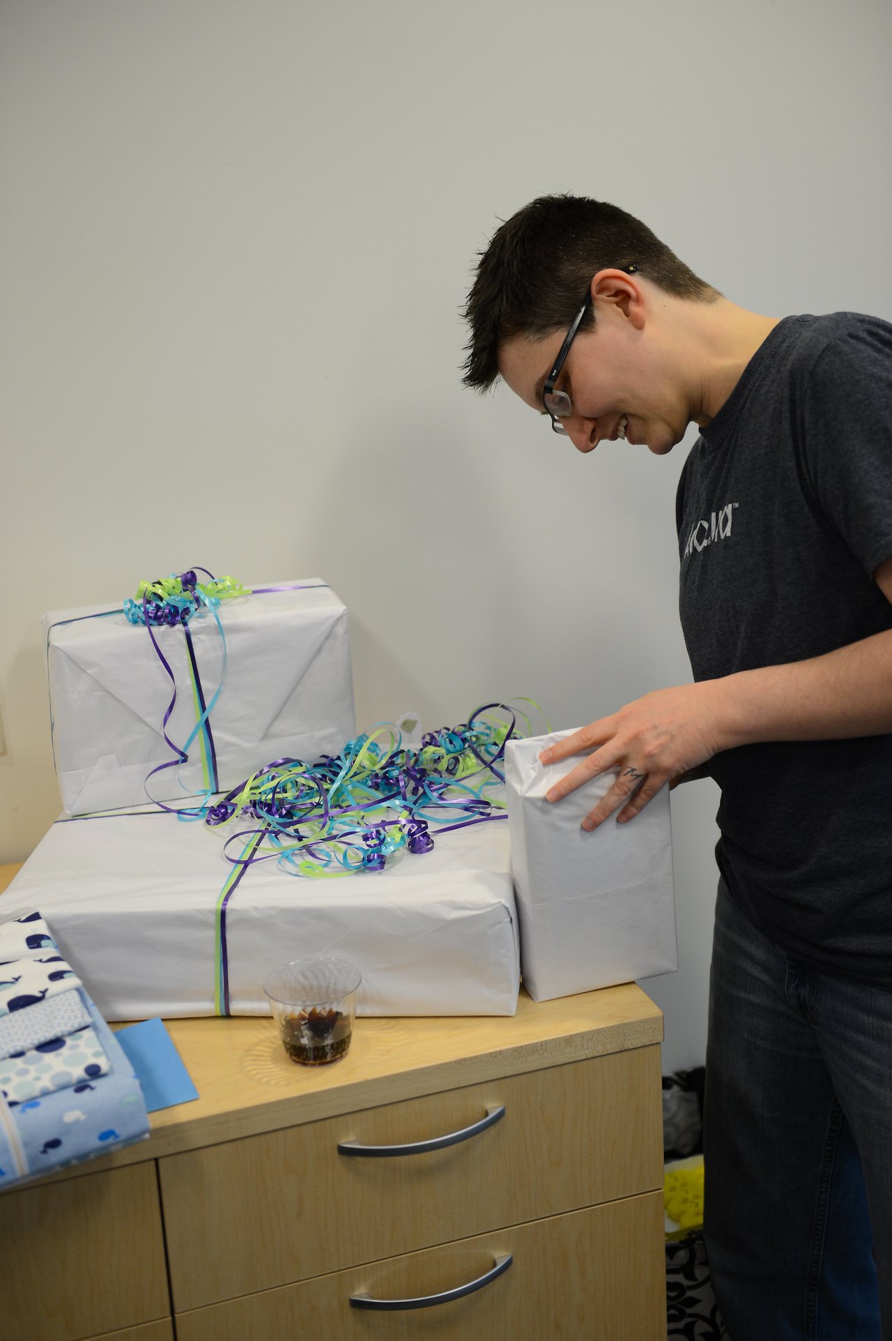 A person in glasses smiles while holding a wrapped gift near a table with more presents.