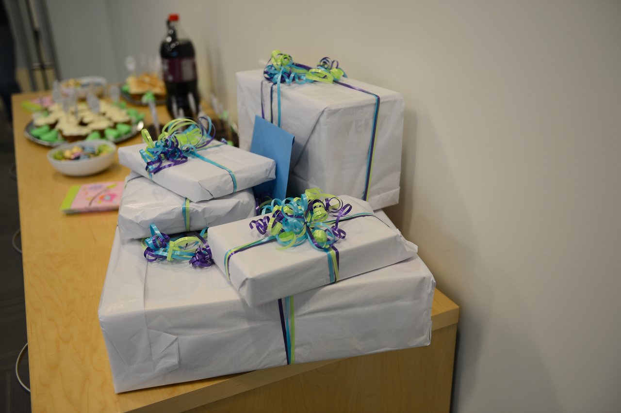 A stack of wrapped gifts with ribbons on a table, with snacks and drinks in the background.