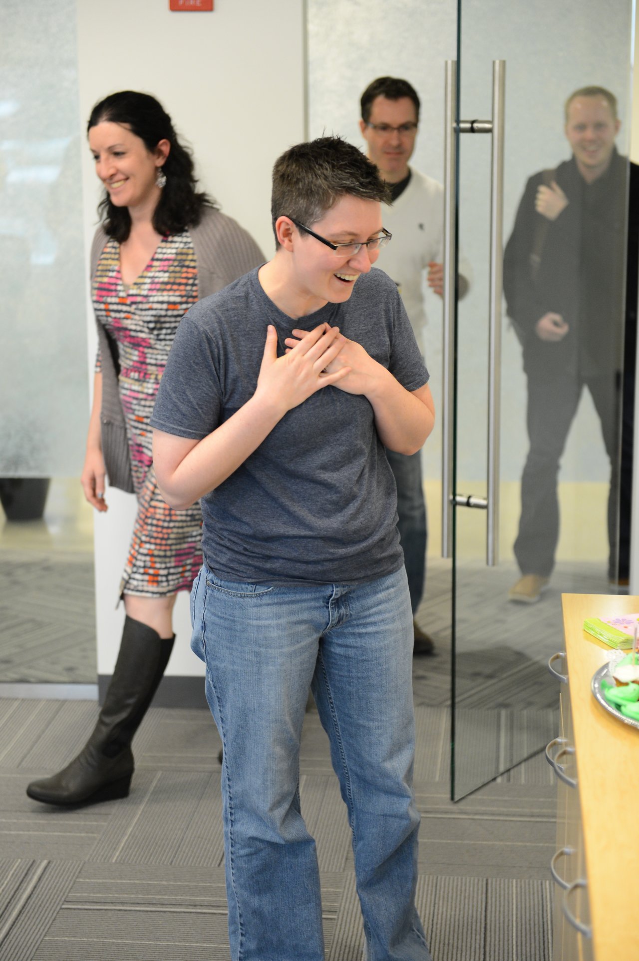 A surprised person enters a room, smiling with hands on their chest, as others follow behind.