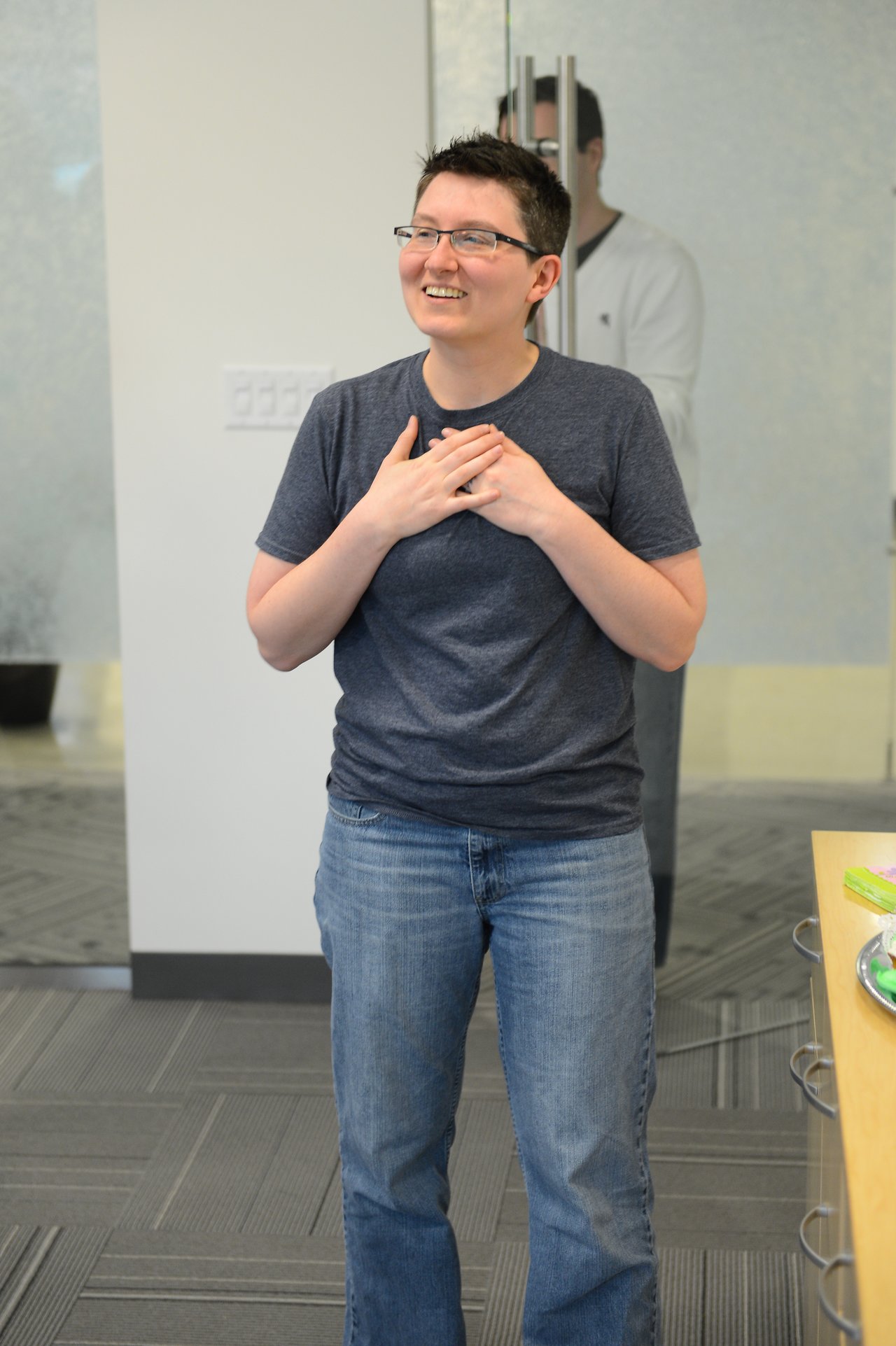 A person in a dark shirt and jeans smiles with hands on their chest, appearing touched or grateful.