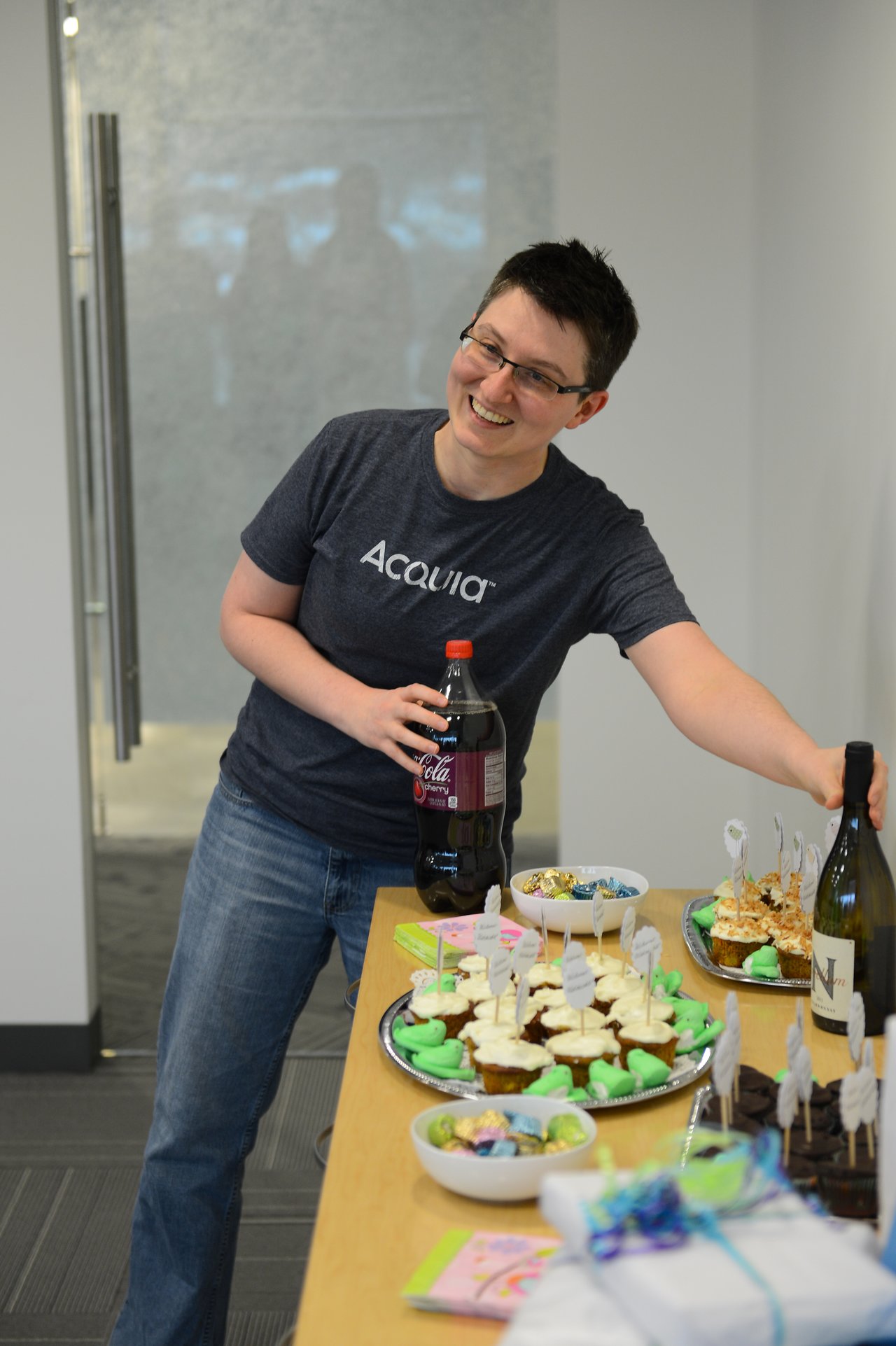 A smiling person in a t-shirt holds a soda bottle while standing by a table with cupcakes and treats.