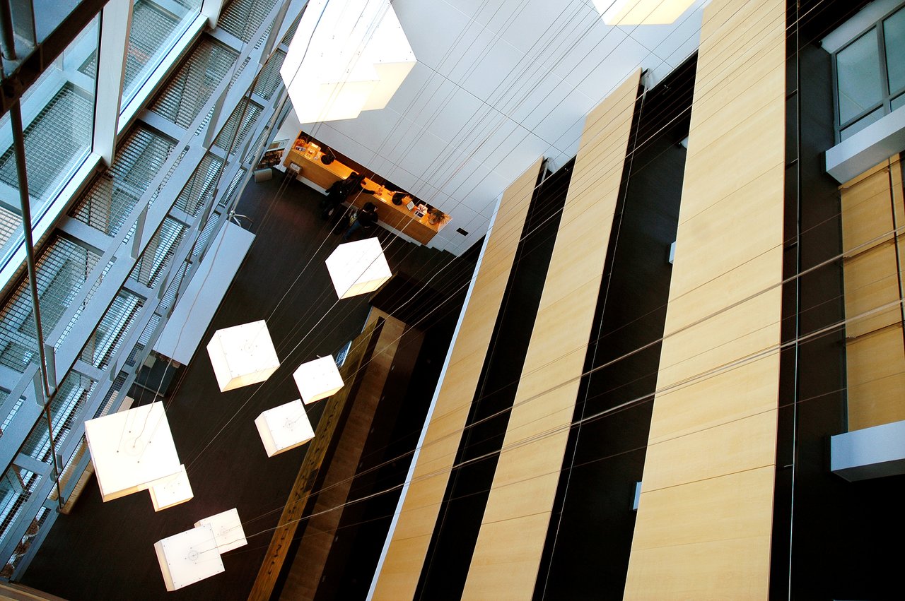 Modern hotel lobby with large hanging cube lights, tall wooden panels, and a reception desk with people checking in.