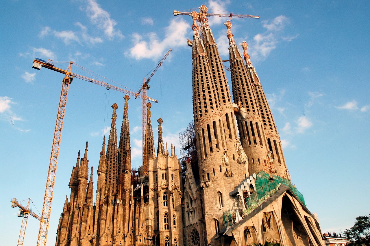 La Sagrada Familia under construction, with several cranes surrounding its tall, detailed towers against a blue sky.