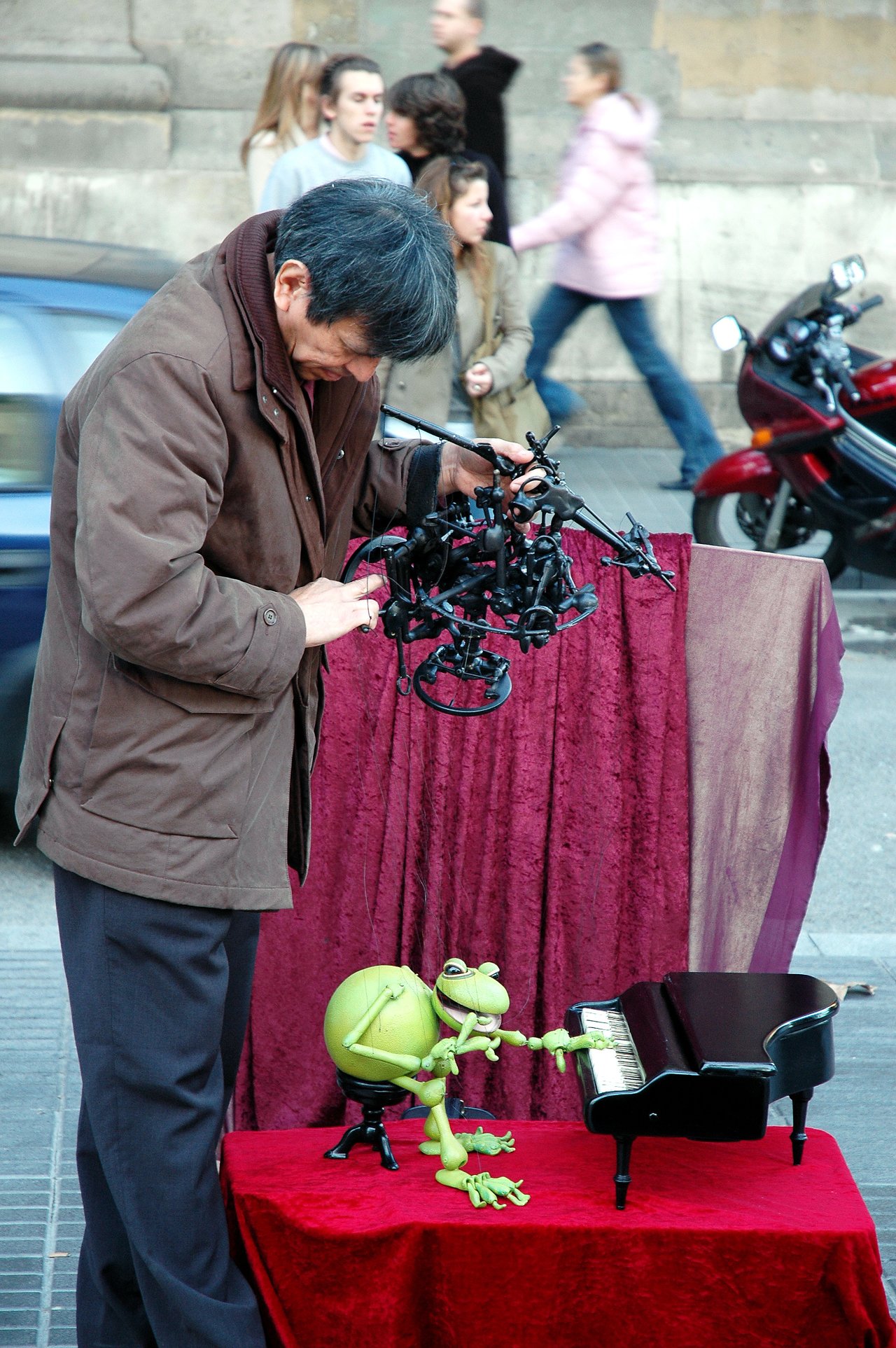 A street performer manipulates a marionette of a frog playing a miniature piano on a red-covered table.