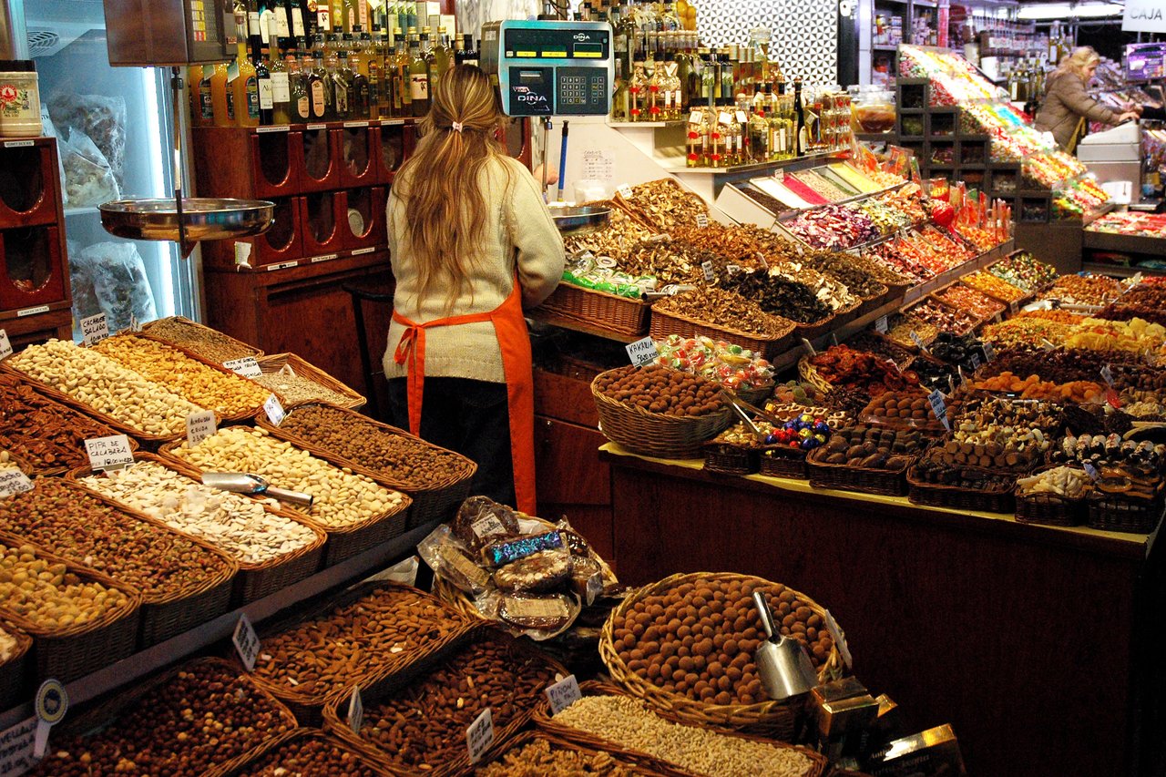 A vendor in an orange apron arranges dried fruits, nuts, and sweets at a busy street market stall.