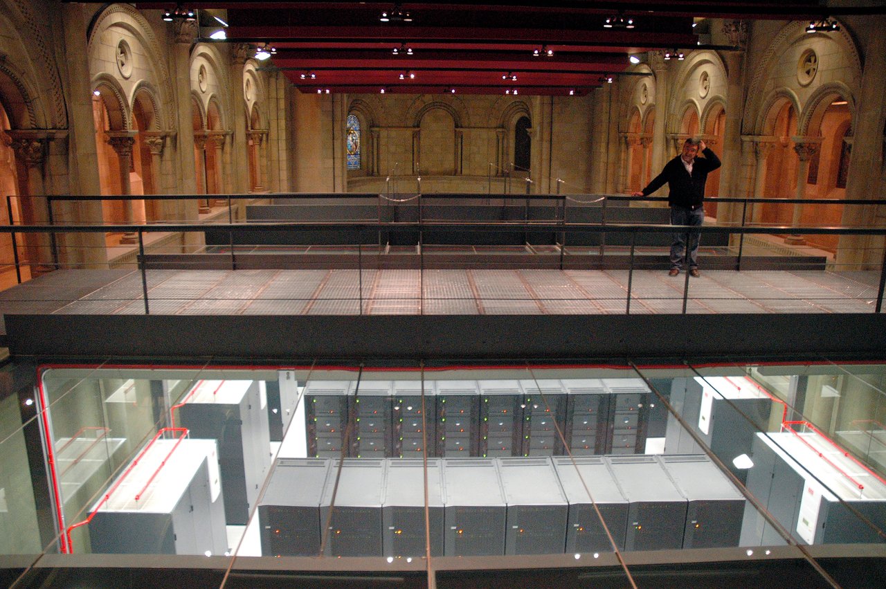 A man stands on a walkway above a large supercomputer housed in a historic building with arched ceilings.