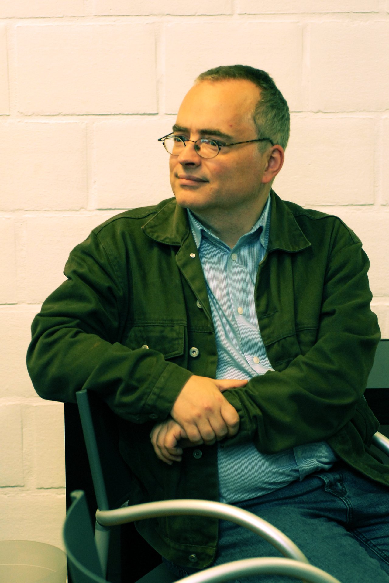 A man wearing glasses and a green jacket sits with his arms crossed, looking to the side.