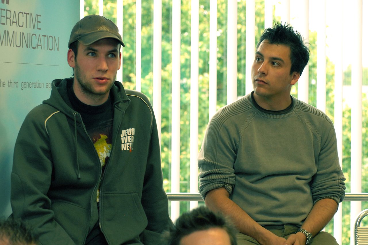 Two men sit indoors near a window, one speaking while the other listens attentively.