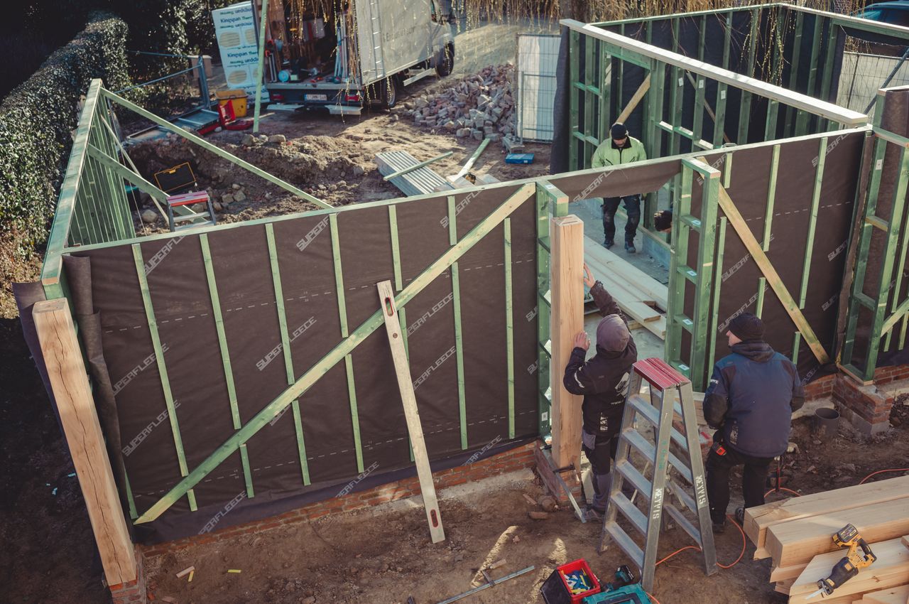 Workers building a wooden frame for what will become a two-car garage. Various tools, including a level and saw, are scattered around.